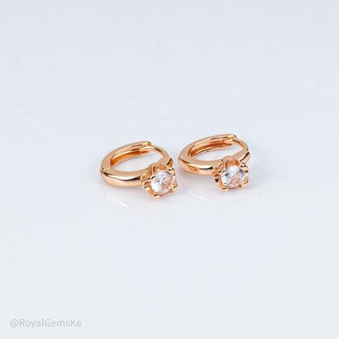 A pair of rose gold huggie hoop earrings, each featuring a small flower-like cluster of clear cubic zirconia stones in a prong setting. The earrings are photographed on a clean, light gray reflective surface.
