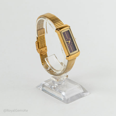 A gold-tone rectangular analog watch with a dark dial and mesh bracelet, displayed on a clear acrylic stand against a light gray background.