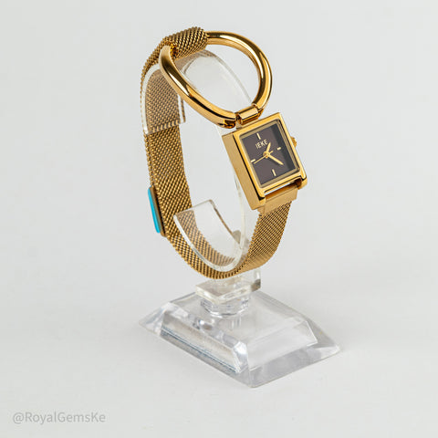 A gold-tone rectangular watch with a black dial and mesh strap, displayed on a clear acrylic stand against a light gray background.