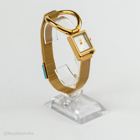 A gold-tone rectangular watch with a white dial and mesh strap, displayed on a clear acrylic stand against a light gray background.
