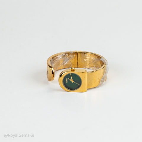 A gold-plated cuff bracelet watch with a green dial and gold hands, partially wrapped in protective plastic on a white background.