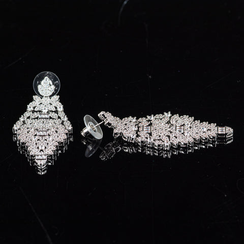 A pair of silver-toned statement earrings covered in clear crystals, displayed on a reflective black surface. One earring is shown upright, revealing its circular clear stud and intricate tiered crystal design, while the other lies horizontally to show the back with its clear silicone stud and screw post.