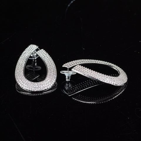 A pair of silver-toned teardrop-shaped hoop earrings paved with clear rhinestones, displayed on a reflective black surface.