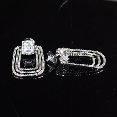 A pair of silver-toned geometric drop earrings with a central clear rectangular gemstone and rows of small clear stones, displayed on a glossy black reflective surface.