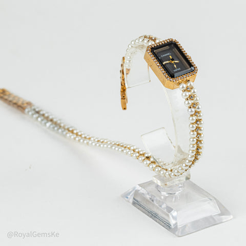 A women's rectangular quartz watch with a gold-tone case, black dial, and a dual-strand bracelet featuring white pearls and gold links, displayed on a clear acrylic stand.