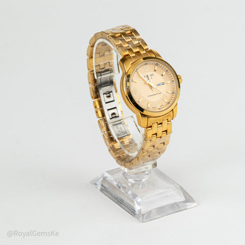 A gold-tone stainless steel watch with a champagne dial and date window, displayed on a clear acrylic stand against a light gray background.