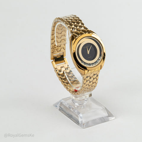 A gold-tone oval watch with a black dial and bezel details, displayed on a clear acrylic stand against a light gray background.