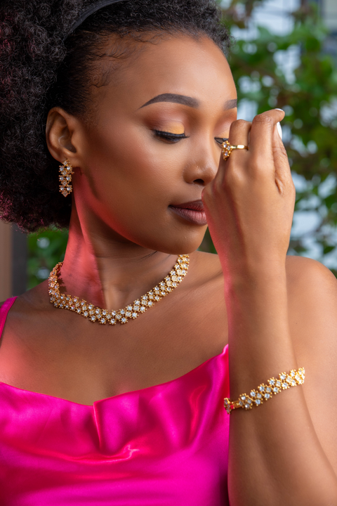 Woman wearing gold jewelry including a necklace, bracelet, and ring, with a blurred outdoor background.