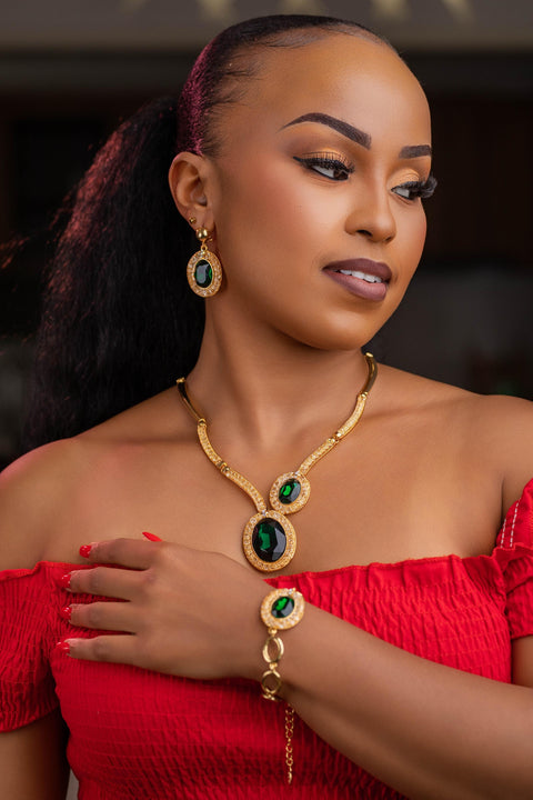 Woman wearing gold jewelry with green gemstones, including a necklace and bracelet, against a dark background.